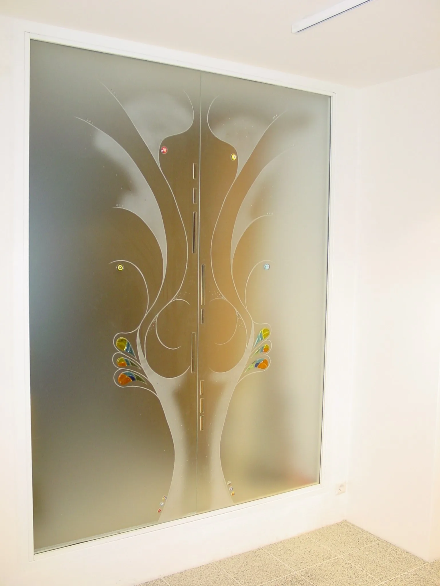Glaselement in einer Wandnische mit sandgestrahltem Baummotiv und farbigen Glasstücken